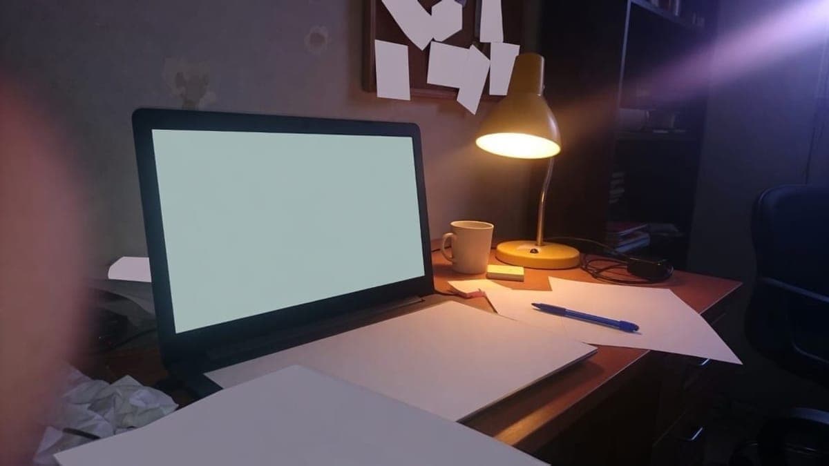 Scrivania di sera con un portatile dallo schermo vuoto, una lampada accesa, una tazza e fogli sparsi.